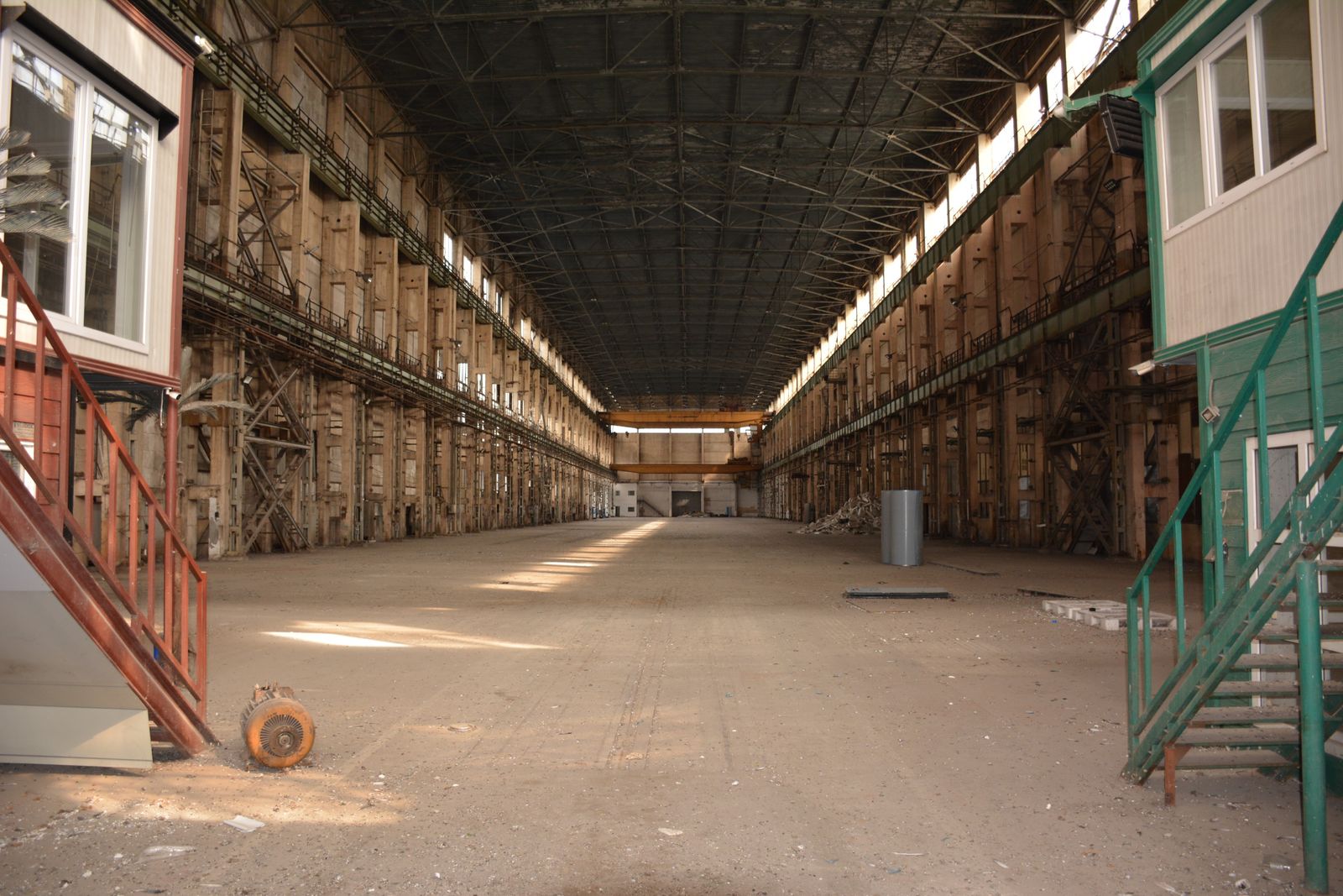 A vast industrial hangar with cathedral-like proportions. The space stretches endlessly, with tall concrete walls, rhythmic columns, and metal structures that create a sense of symmetry and infinity.